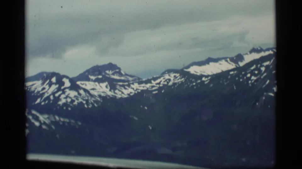1977 Alaska 8mm aerial view of a snow-streaked mountain range from an aircraft, with soft focus and gray cloud cover.