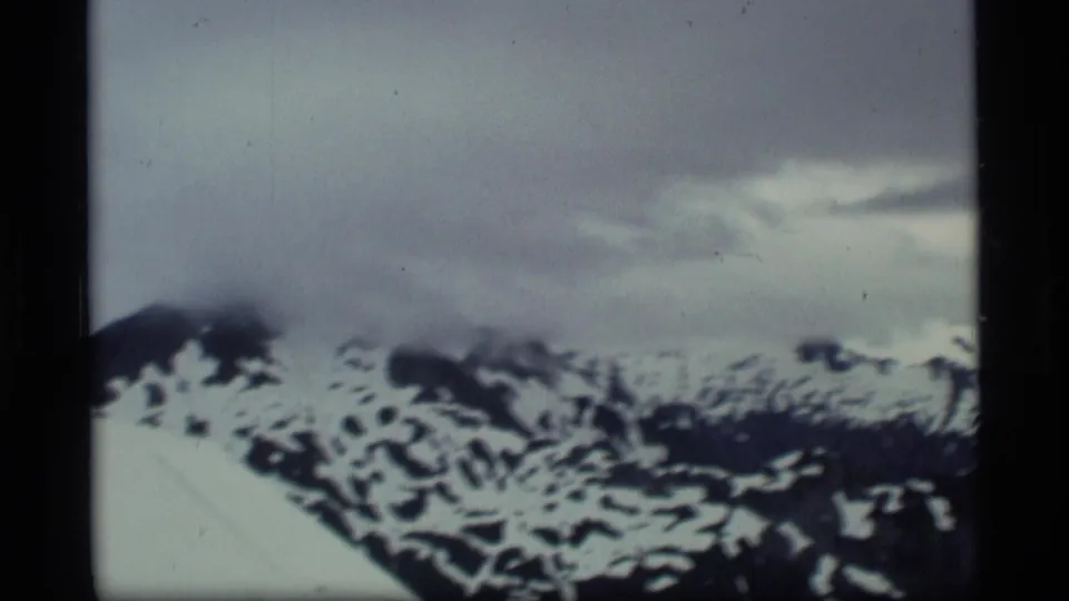 1977 Alaska aerial frame with a partial aircraft wing edge, blurred snow-covered mountain ridges, and low cloud cover in 8mm film texture.