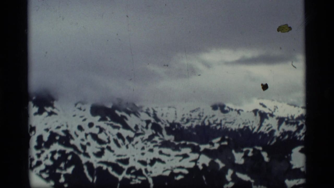 1977 Alaska 8mm frame showing a distant snow-streaked mountain ridge beneath heavy cloud cover and visible film wear.