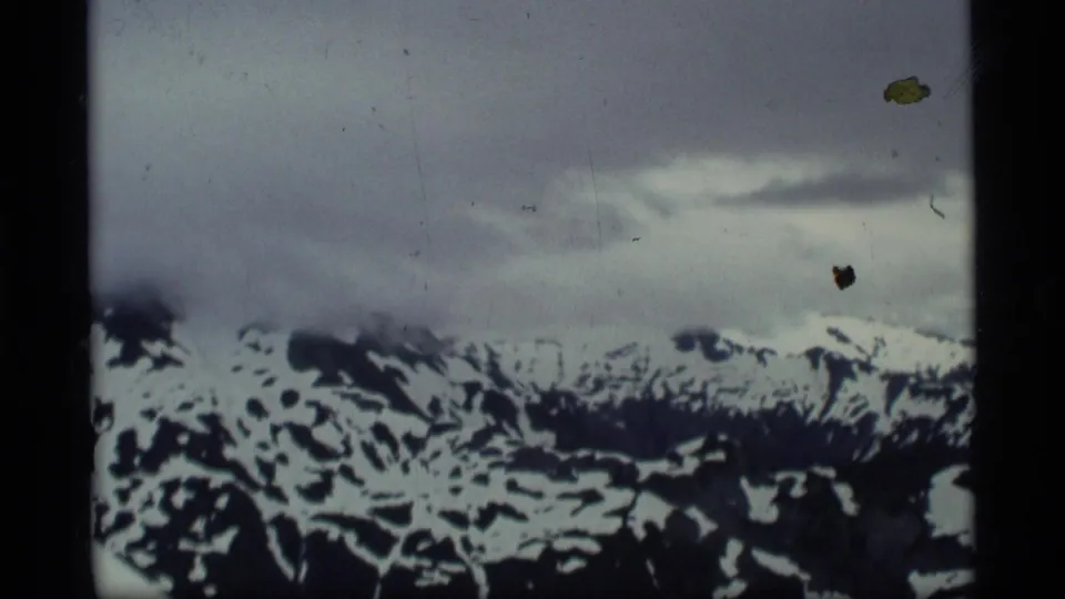 1977 Alaska 8mm frame showing a distant snow-streaked mountain ridge beneath heavy cloud cover and visible film wear.