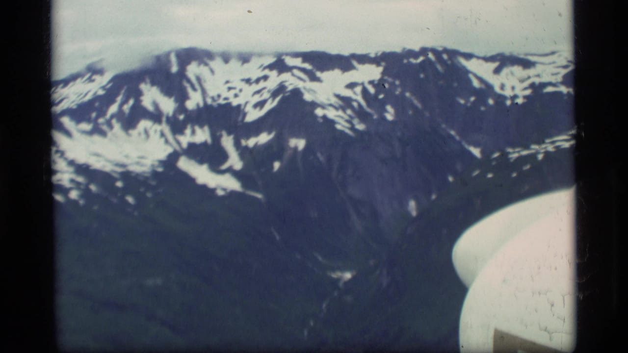 Passenger-view 8mm frame over Alaska, with snow-capped mountains and the curved edge of an aircraft window or structure.