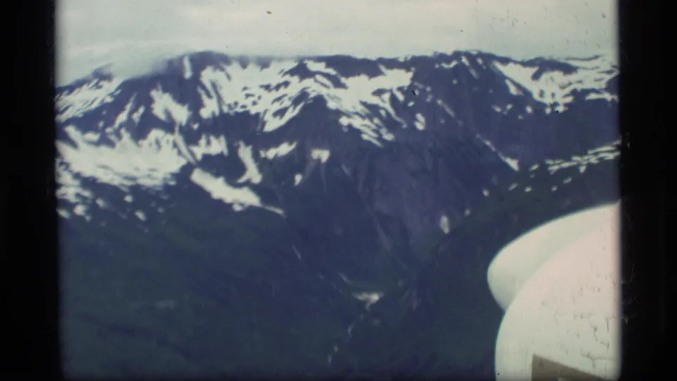 Passenger-view 8mm frame over Alaska, with snow-capped mountains and the curved edge of an aircraft window or structure.