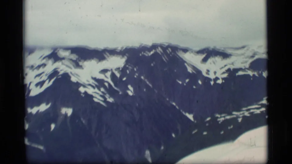 1977 Alaska home-movie frame of rugged mountains and snow patches, filmed from an aircraft with part of the plane visible.