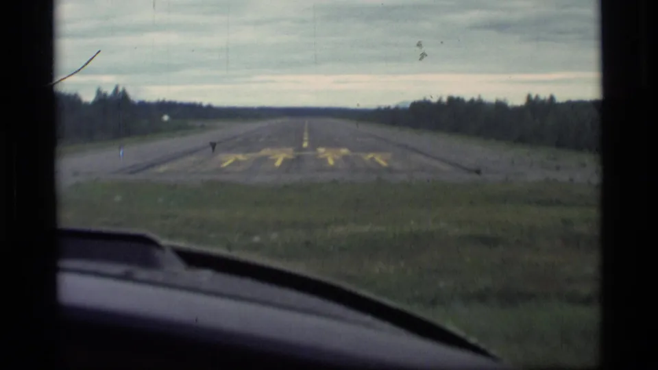 Aircraft approach frame in Alaska, showing a runway through the windshield with grassy shoulders and distant trees.
