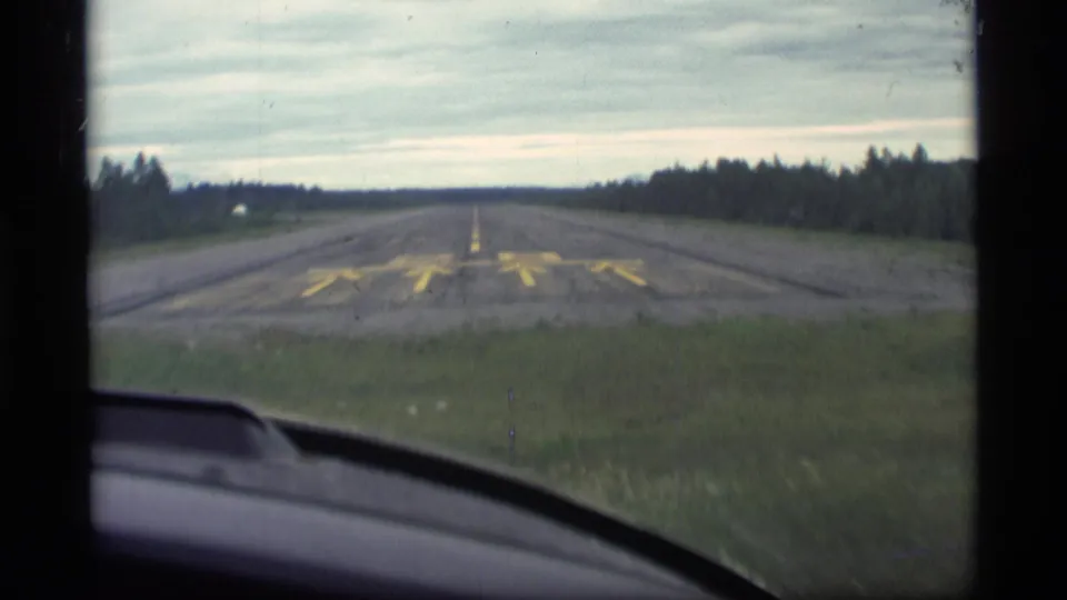 1977 Alaska home-movie frame showing Runway 28 ahead through a windshield, bordered by dark forest under an overcast sky.