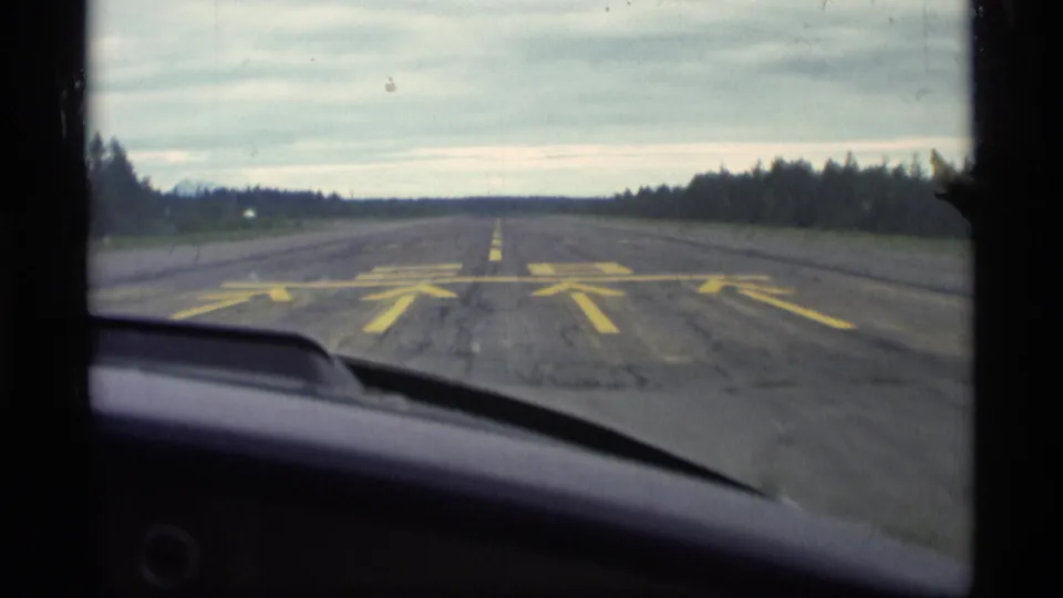 Windshield view of a paved rural Alaska runway with yellow markings, a dashboard edge, and trees along the horizon.