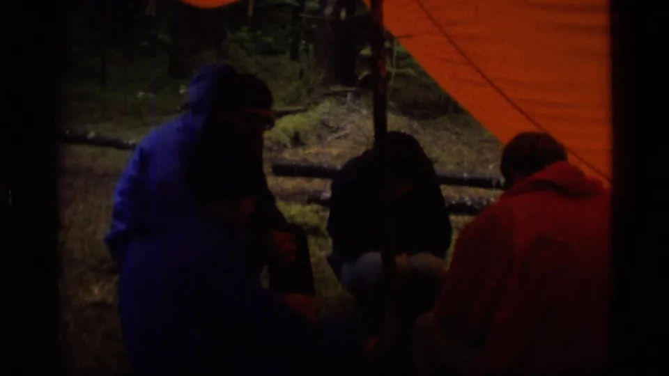 1977 Alaska campsite frame with several people under an orange tarp, partly shadowed by trees and film exposure.