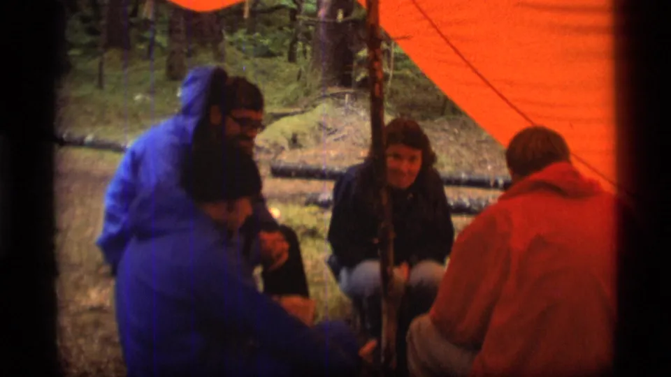 Home-movie view of adults gathered under an orange tarp at a wooded Alaska campsite, with faces partly visible.