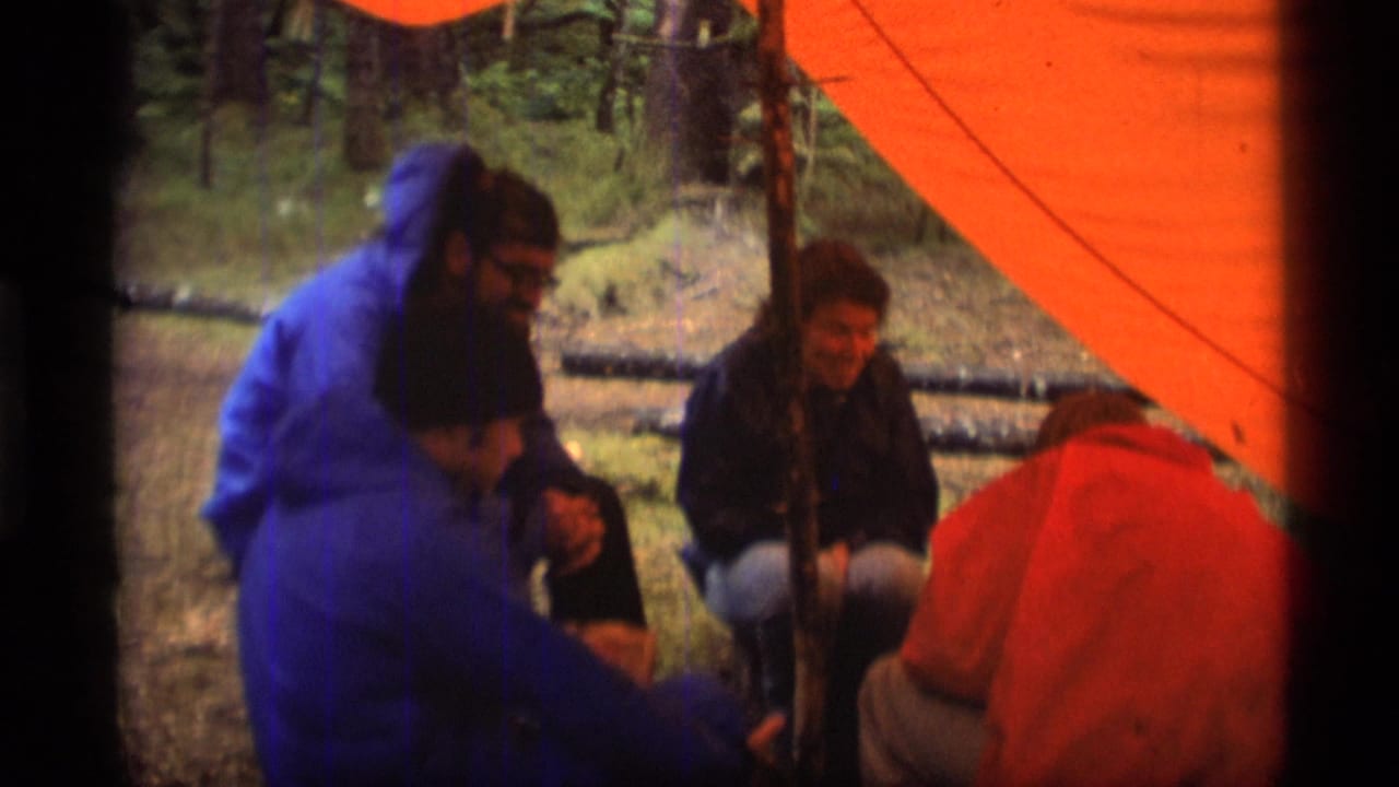 Campers in rain gear sit under a bright orange tent in a forest setting.