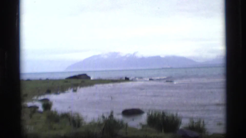 1977 Alaska coastal view with calm water, low shoreline, and distant mountains partly softened by haze.