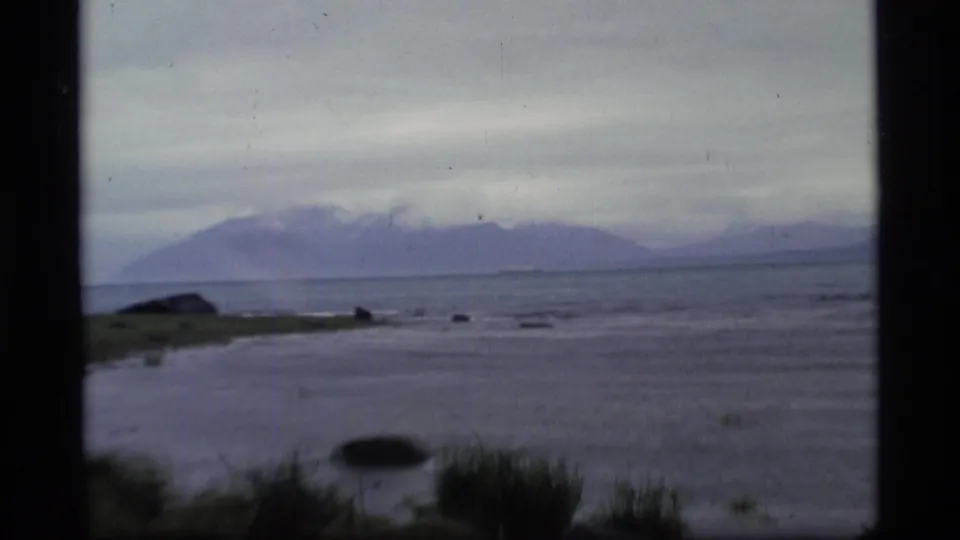 1977 Alaska home-movie view of rocky shoreline water with low clouds over distant mountains.