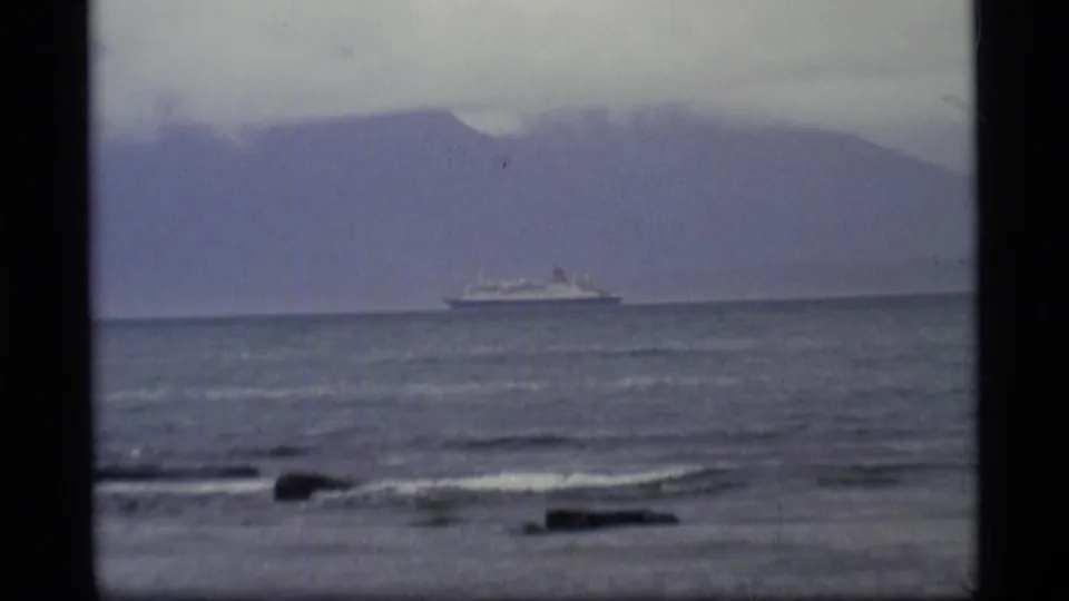 1977 Alaska coastal frame showing a large ship moving across water in front of hazy mountains.