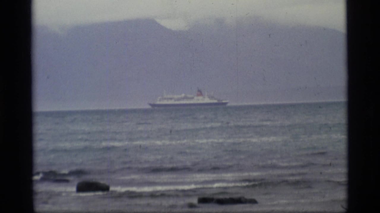 Distant passenger ship crossing grey water with rocks and mountains beyond.