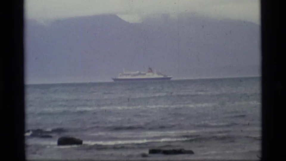 1977 Alaska 8mm frame showing a distant passenger ship offshore, low rocks in the foreground, and hazy mountains under an overcast sky.