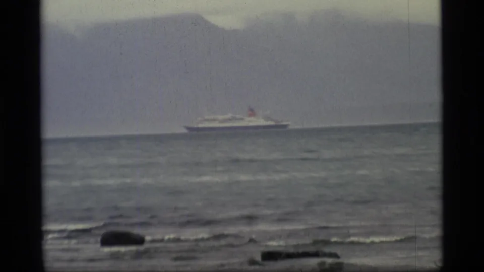 1977 Alaska home-movie frame with a distant passenger vessel on grey water, shoreline rocks below, and a muted mountain backdrop.