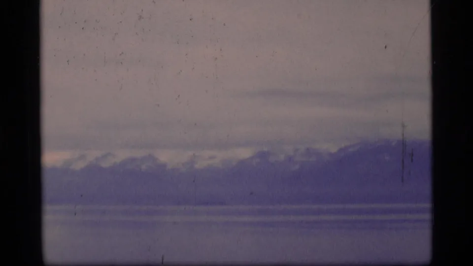 1977 Alaska 8mm frame shifting from the ship to a wide view of water, distant mountains, snow patches, and a subdued cloudy sky.