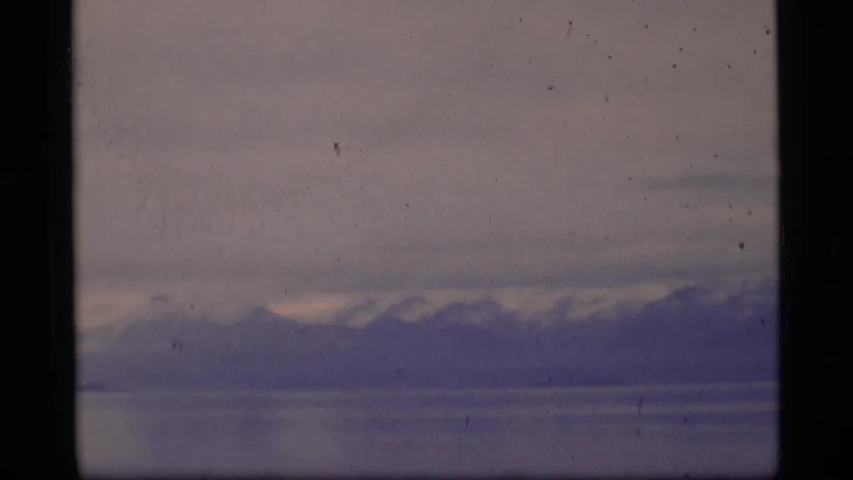 1977 Alaska home-movie frame showing a soft distant mountain range across water, with grain, dust, and muted purple-blue color shifts.