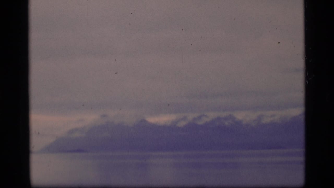 Cloudy sky over distant mountains and calm water or ice, framed by dark film borders.
