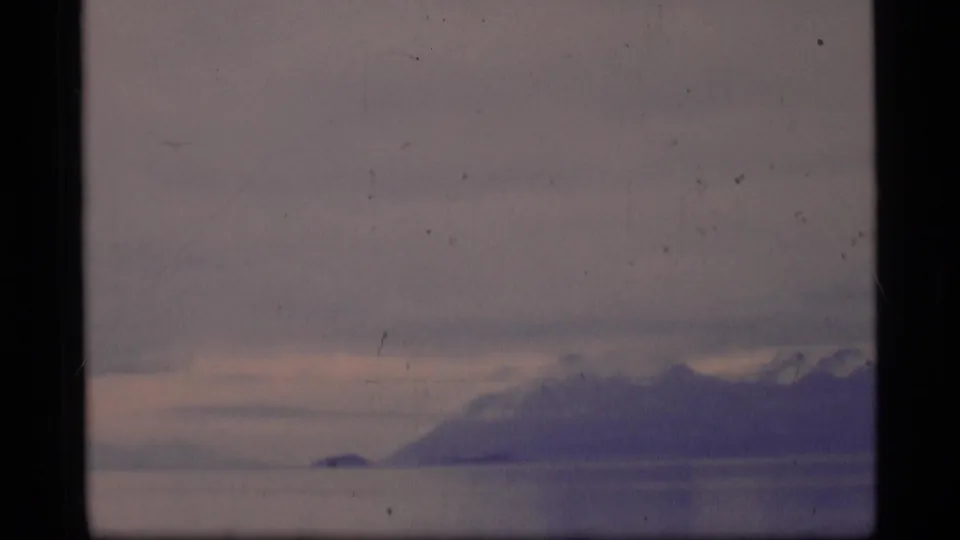 1977 Alaska 8mm view of a snowy mountain range beyond a quiet water or ice surface beneath a low, overcast sky.