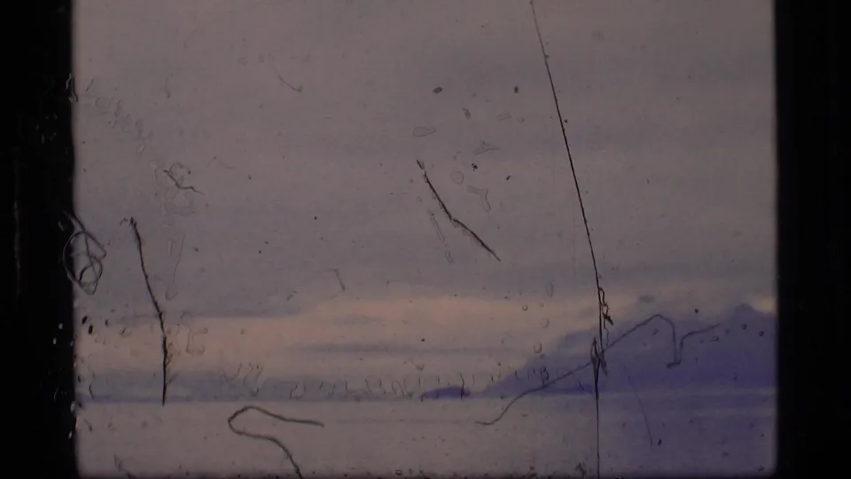 Faded 1977 Alaska 8mm frame with distant mountains across water or ice, a low cloud band, and prominent surface scratches.