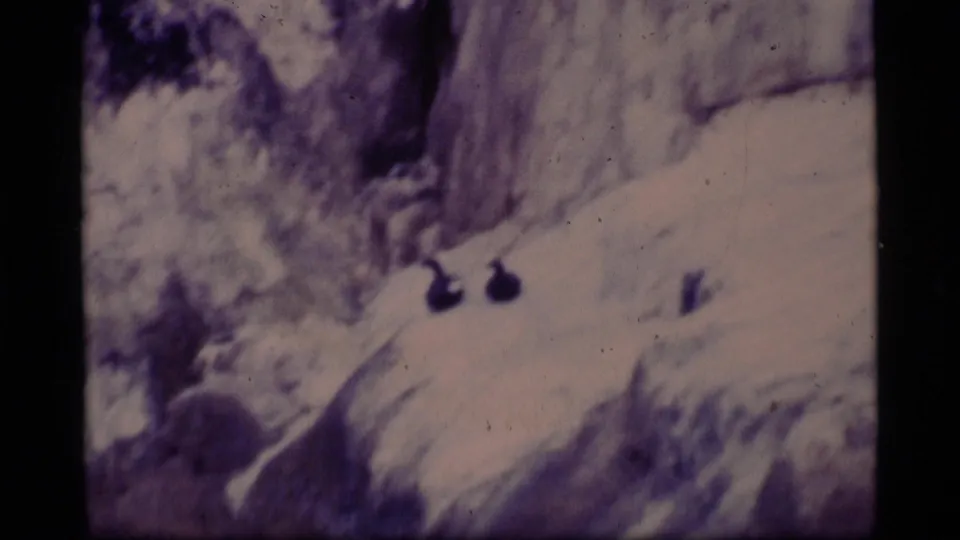 1977 Alaska 8mm frame of a snow-covered ledge or slope with rocky terrain and dark indistinct forms, rendered in soft grain and purple color shift.