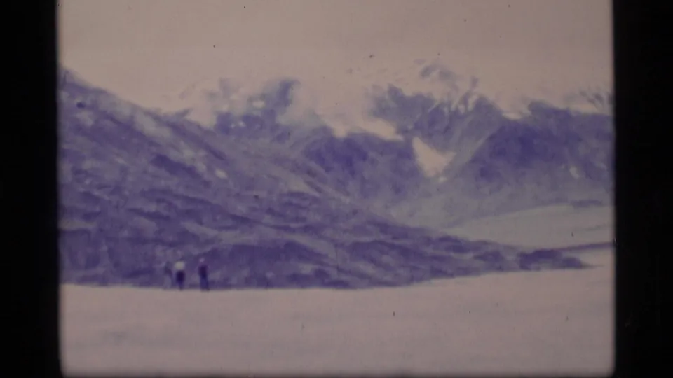 1977 Alaska 8mm frame showing two distant figures on a snowfield beneath broad mountain ridges and a pale sky.