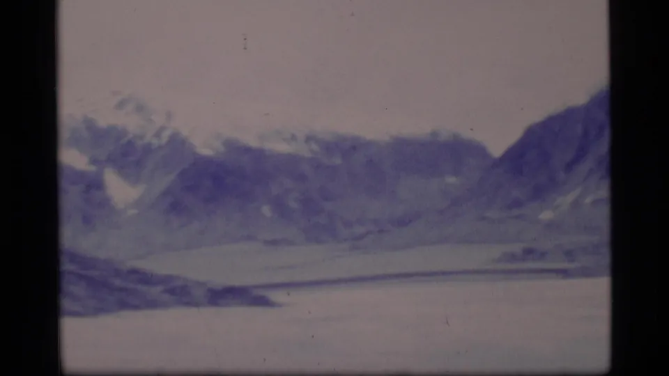 1977 8mm Alaska landscape with distant snow-dusted mountains, a pale frozen lake, and visible film gate edges.