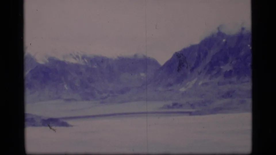Soft 1977 8mm view of an Alaskan mountain ridge across a frozen lake, with muted color and film debris visible.