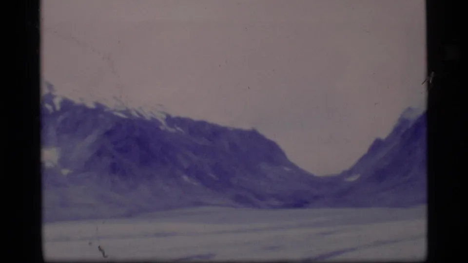 1977 8mm Alaska frame showing a broad mountain slope above a frozen lake, with violet color shift and visible grain.
