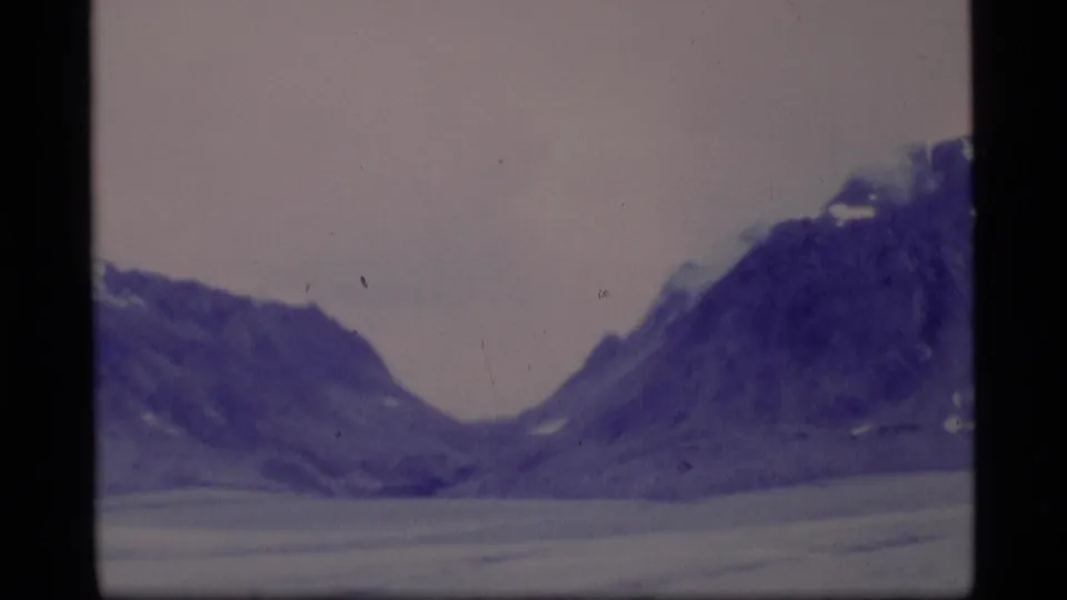 Soft 1977 8mm Alaska landscape with mountains forming a valley above a pale frozen lake and muted sky.