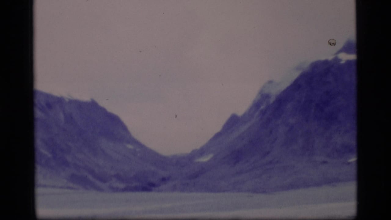 1977 Alaska 8mm frame showing rugged mountain slopes around a broad valley or waterline under an overcast sky.