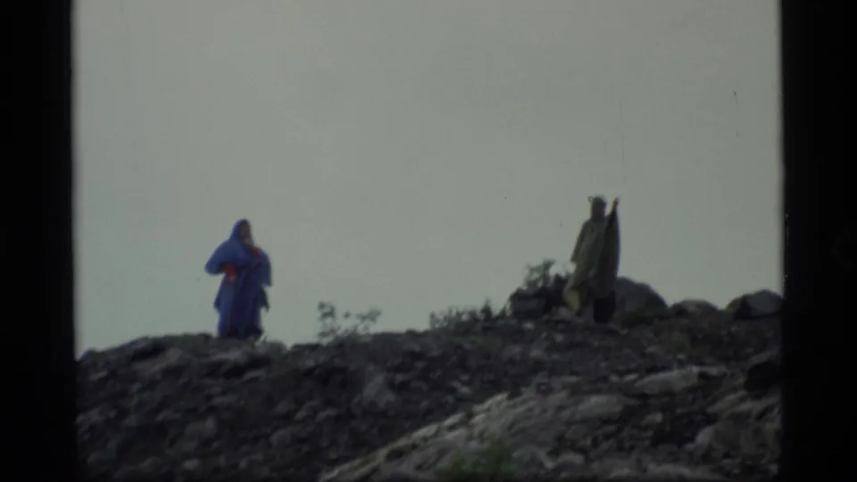 1977 Alaska 8mm frame showing two distant travelers on rocky ground, one in blue rain gear and another darker figure near low shrubs.
