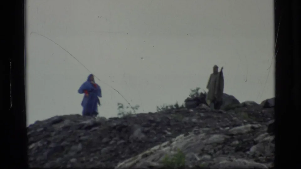 1977 Alaska 8mm frame of two distant people on a rocky overlook, with muted colors, low shrubs, and an overcast sky.