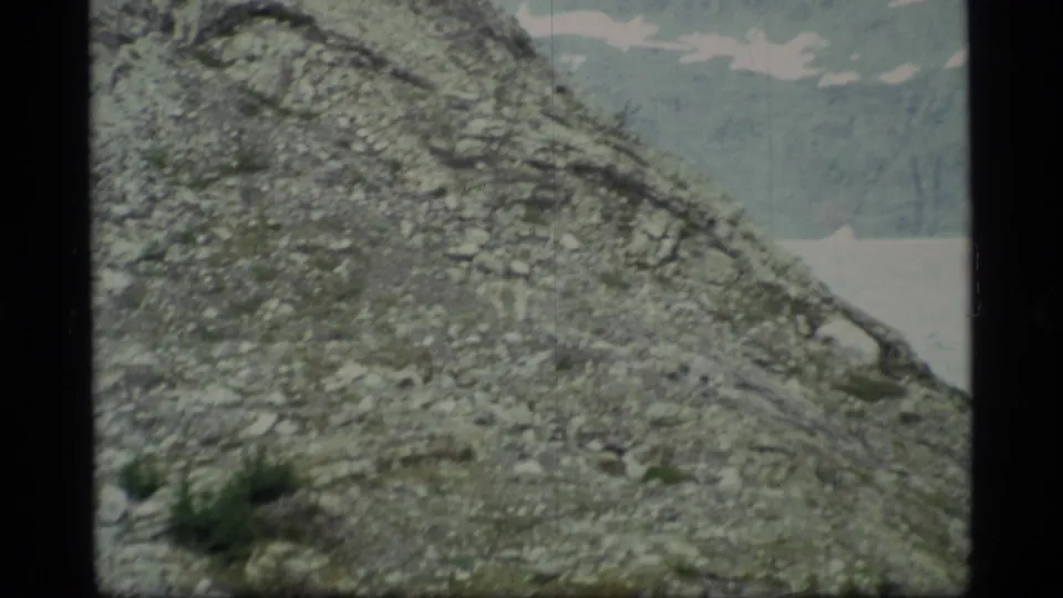 Home-movie view from 1977 Alaska showing a stony mountainside, sparse green growth, and a light-toned body of water in the distance.