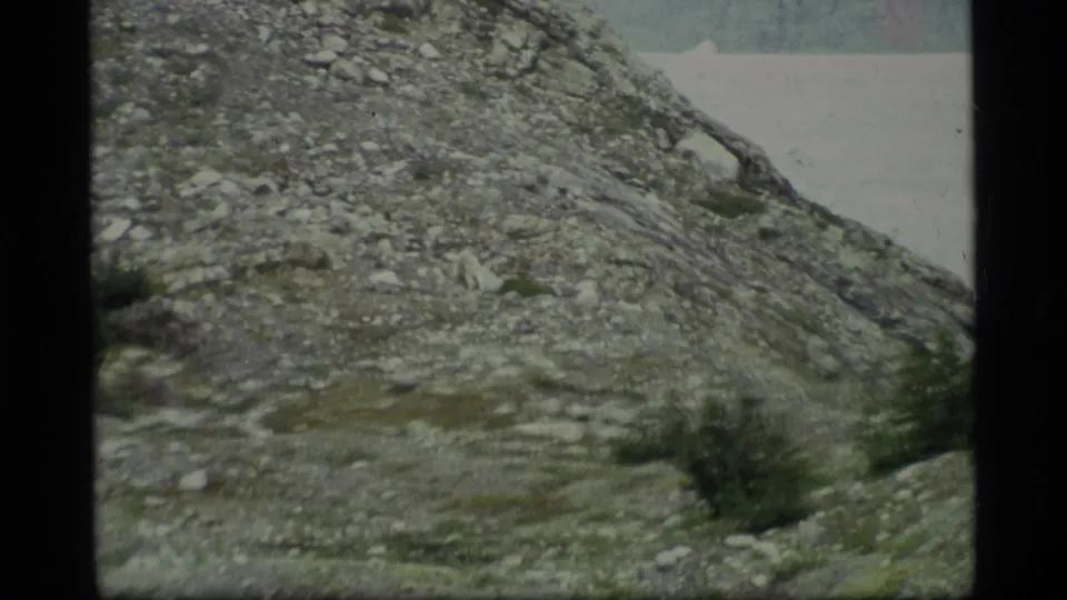 1977 8mm Alaska frame showing a rocky mountain slope descending toward distant pale water, with sparse vegetation in the foreground.