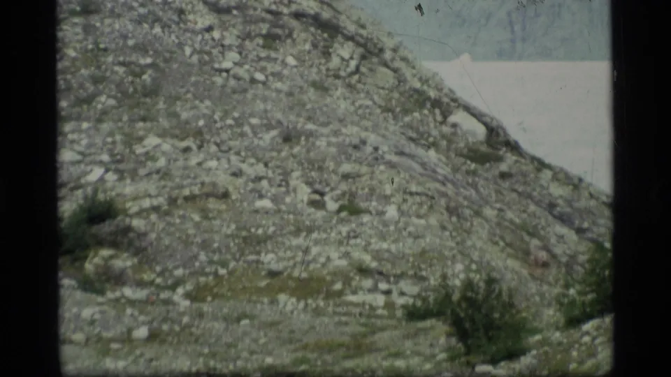 1977 Alaska 8mm frame of a rock-strewn hillside, sparse vegetation, and a pale horizon behind the sloping terrain.
