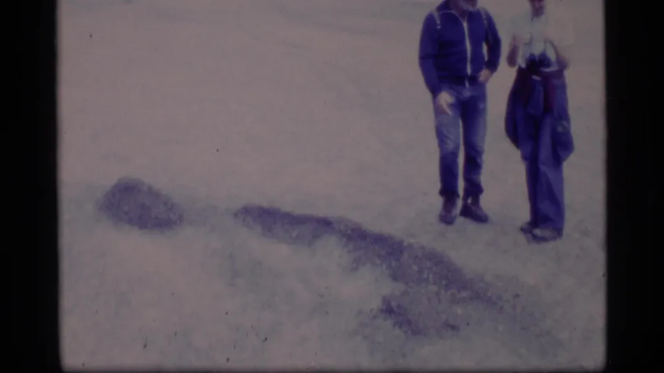 1977 Alaska 8mm frame showing two adult men standing on pale ground near the base of a rocky hillside, with faded color and soft focus.