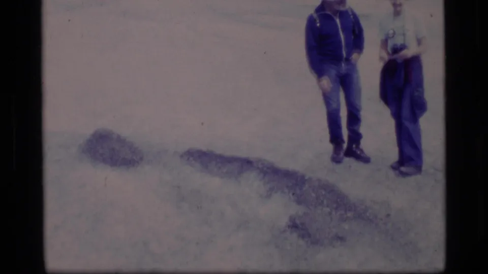 1977 Alaska home-movie frame showing two adults in winter clothing standing on a frozen surface near dark patches of snow or ice.