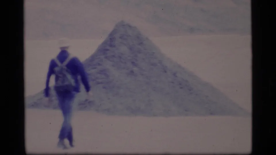 1977 Alaska frame of a person in a blue jacket and backpack walking across a pale frozen landscape toward a conical snow or ice mound.