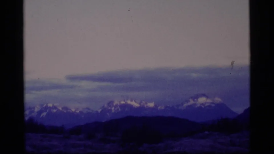 1977 Alaska home-movie frame with distant snow-capped mountains under a pale sky and dark low hills in the foreground.