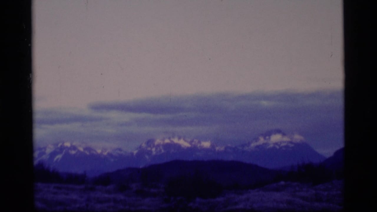 1977 Alaska 8mm frame showing distant snow-capped mountains, dark foreground hills, meadow, and a soft lavender sky.