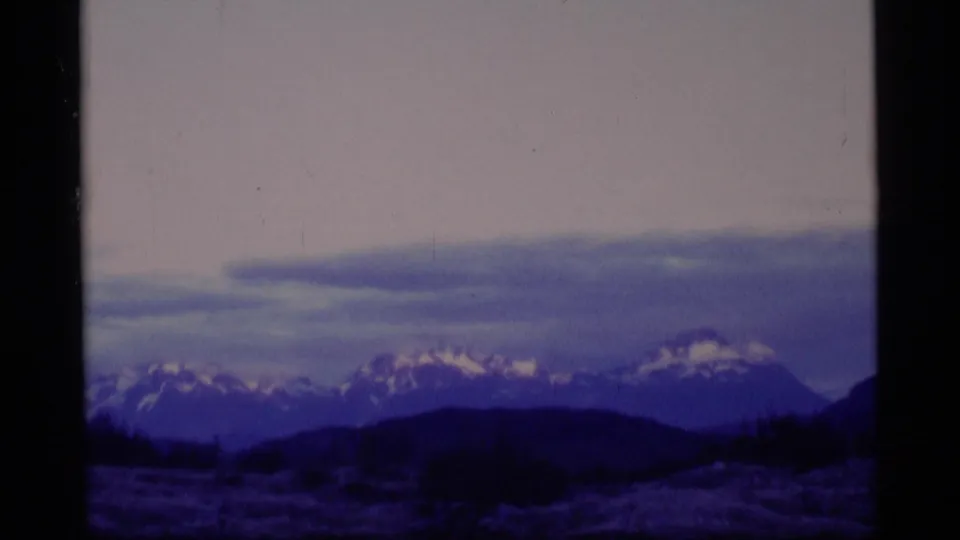 1977 Alaska 8mm frame showing distant snow-capped mountains, dark foreground hills, meadow, and a soft lavender sky.