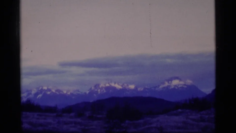 1977 Alaska 8mm frame with snow-dusted peaks, a dark ridge line, low meadow, and muted dusk sky tones.