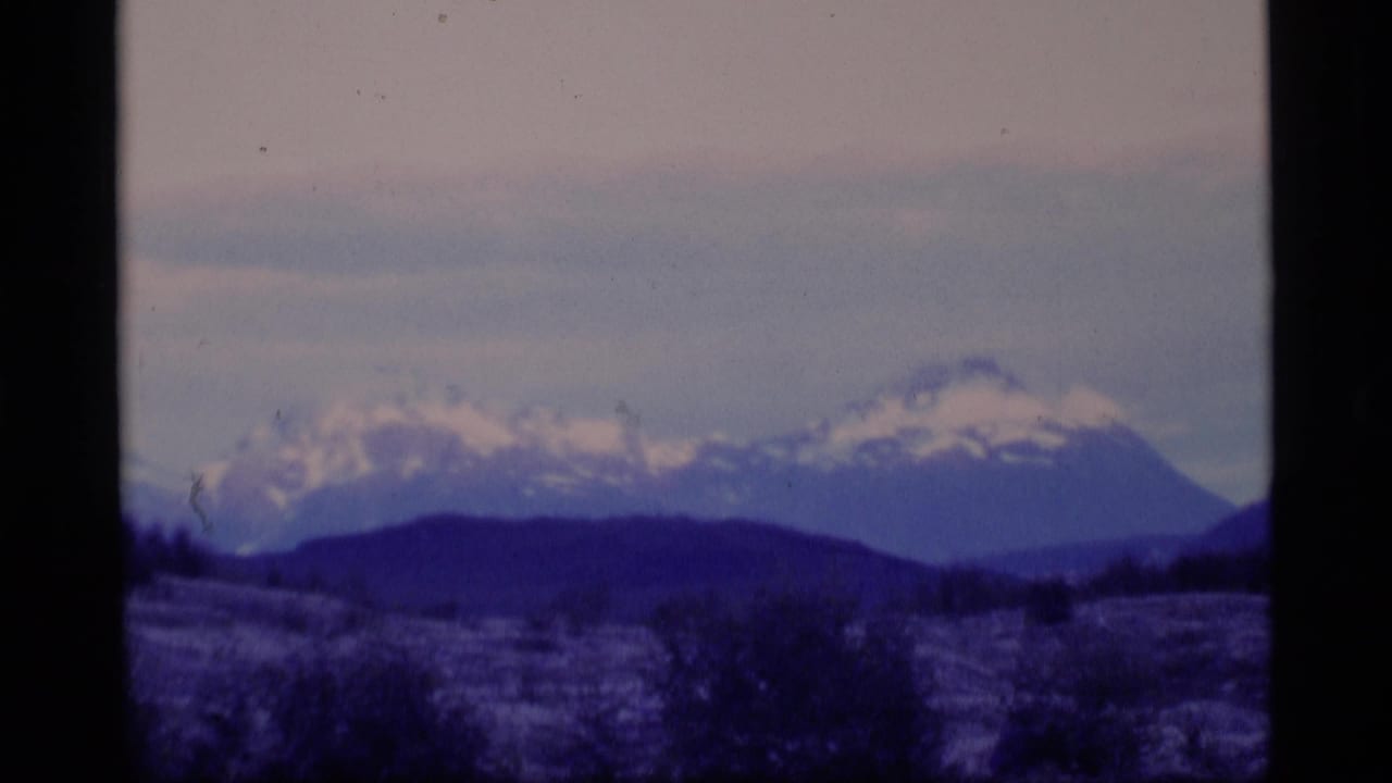 1977 Alaska 8mm frame showing distant snow-capped mountains, dark foreground hills, and a muted overcast sky.