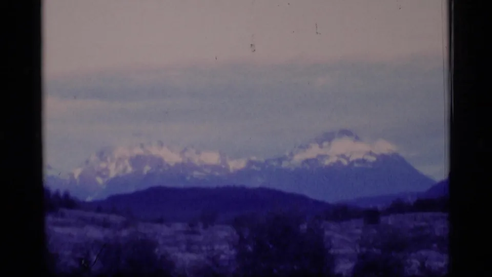Alaska home-movie frame with a snowy mountain range, dark foothills, muted sky, and visible 8mm frame edges.