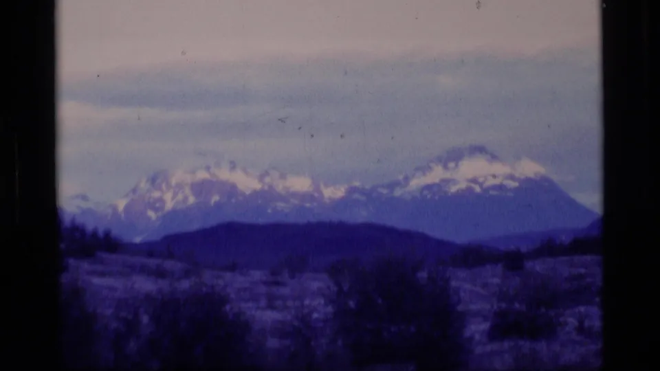 1977 Alaska 8mm landscape frame with snow-topped peaks, dark brush or low hills, and a blue-purple color cast.