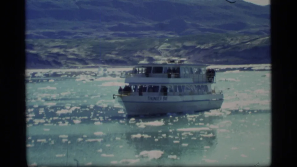 1977 Alaska 8mm frame showing a white tour boat, visible passengers, floating ice, and mountain slopes in the background.
