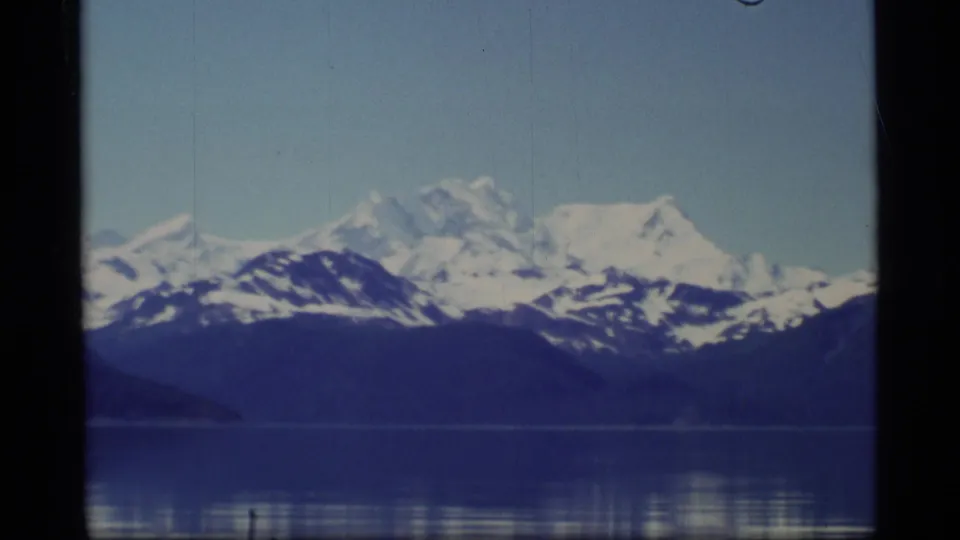 1977 Alaska scenic frame with snow-covered mountains, dark shoreline, and calm blue water under clear sky.