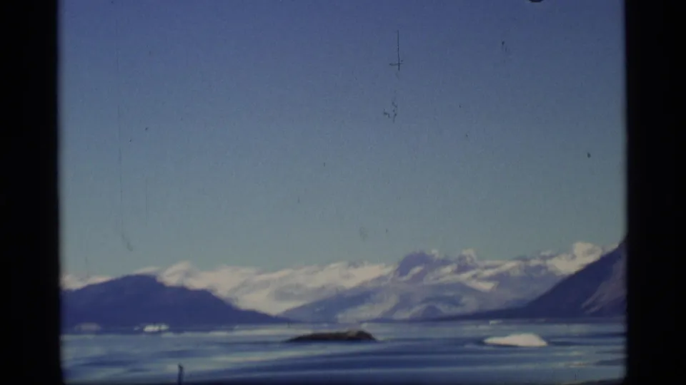 1977 Alaska 8mm view across a calm icy bay toward low landforms and distant snow-covered mountain peaks.