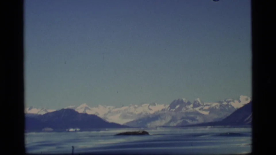 1977 Alaska home-movie frame looking across calm water toward snow-covered mountains and scattered ice in the bay.
